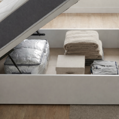 Vista lateral de perfil del canapé abatible de madera blanca Americ abierto, mostrando la profundidad y gran capacidad de su cajón de almacenaje. El interior está organizado con una bolsa de vacío grande para edredones, una pila alta de mantas y una caja organizadora bajo el colchón elevado.