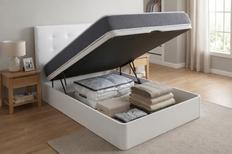Canapé abatible de madera blanca "Americ" abierto en un dormitorio, mostrando su gran capacidad de almacenaje interior con mantas, ropa de cama y cajas organizadas. El colchón está elevado por los pistones hidráulicos. Ideal para ganar espacio.